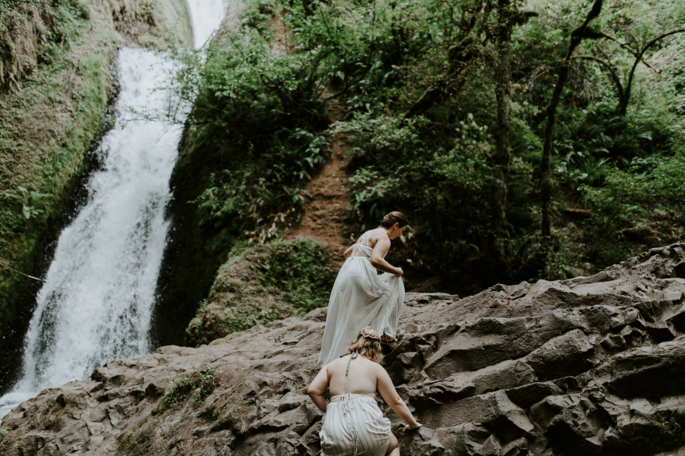 Kate and Audrey climb up a large boulder in front of Bridal Veil Falls. Elopement wedding photography at Bridal Veil Falls by Sienna Plus Josh.