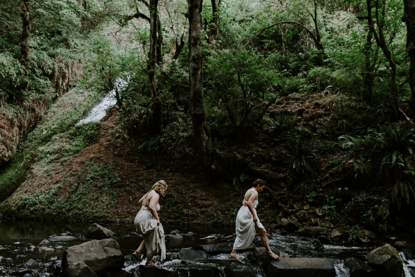 Kate and Audrey crossing a river. Elopement wedding photography at Bridal Veil Falls by Sienna Plus Josh.