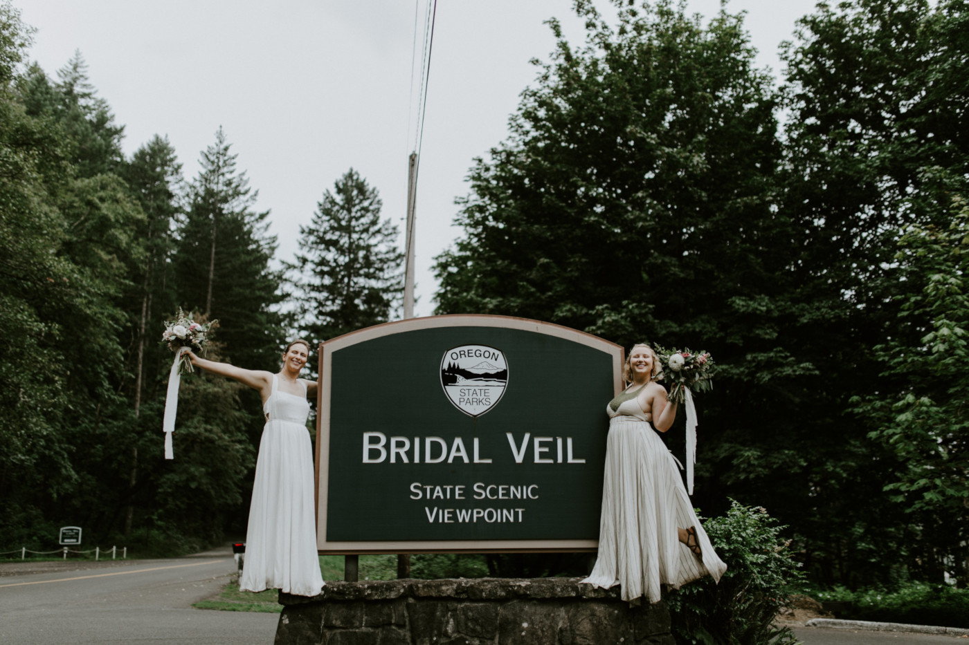 Kate and Audrey stand next to the Bridal Veil Falls sign. Elopement wedding photography at Bridal Veil Falls by Sienna Plus Josh.