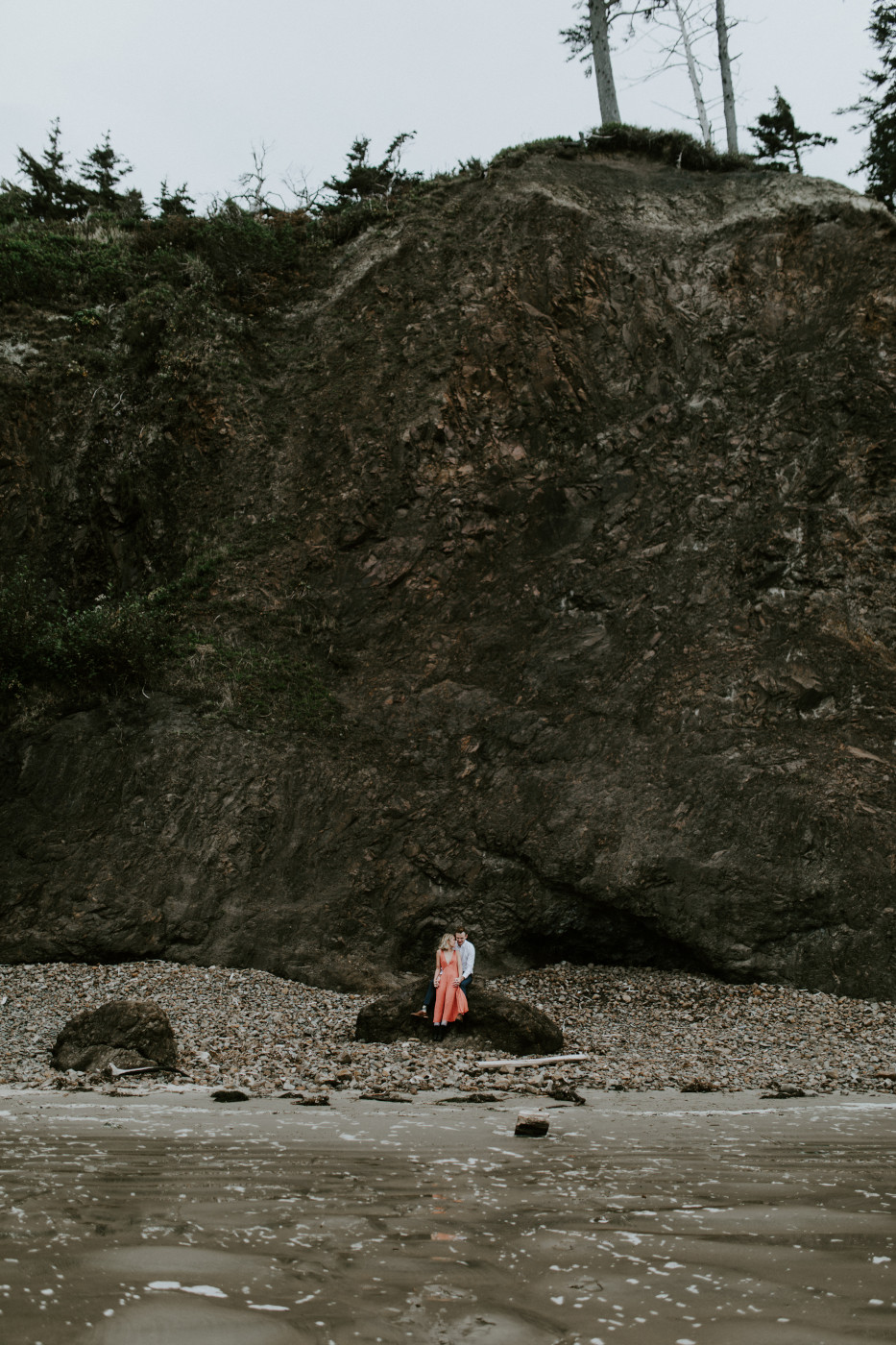 Chelsey and Billy sit together in front of a large cliff. Elopement wedding photography at Cannon Beach by Sienna Plus Josh.