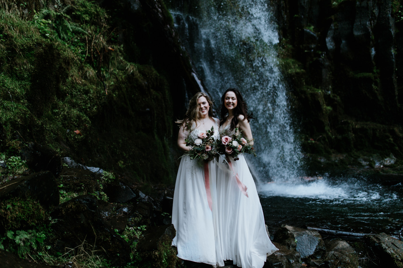 Hayley and Tiffany stand side by side in front of the waterfall. Elopement photography at the Columbia River Gorge by Sienna Plus Josh.