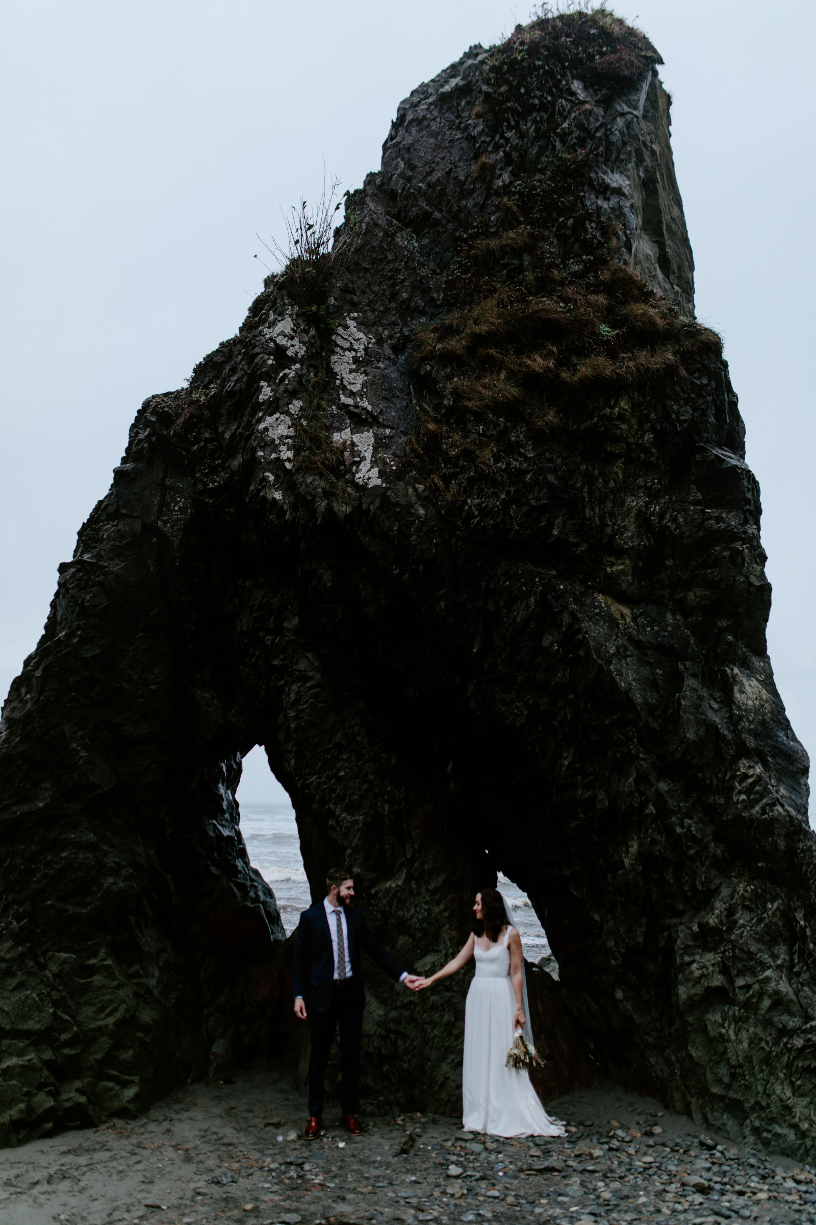 Mollie and Corey stand in front of a large rock on the beach. Elopement photography at Olympic National Park by Sienna Plus Josh.