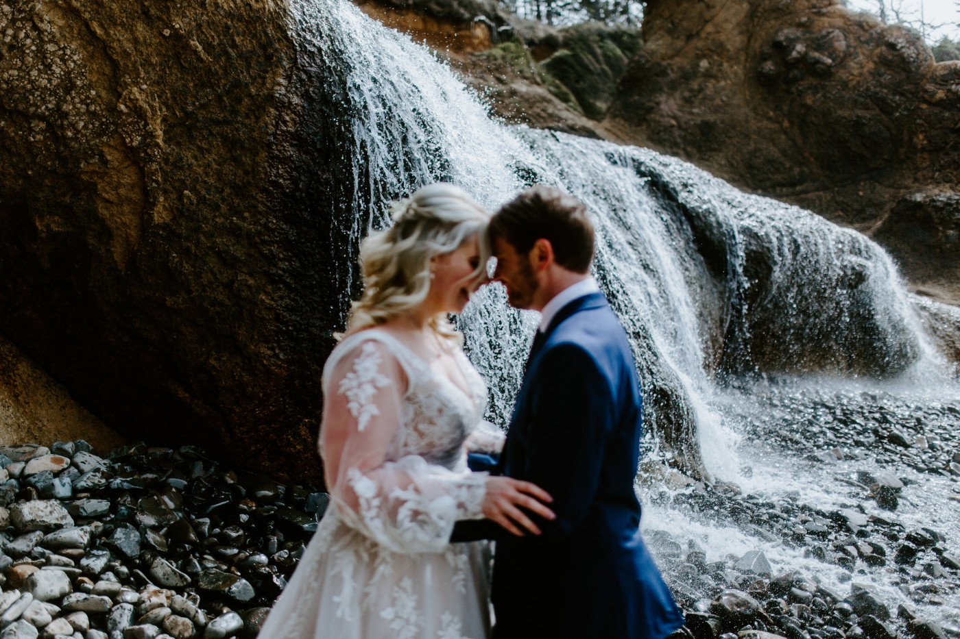 Ben and Katie stand in front of a waterfall at the Oregon coast on the beach.