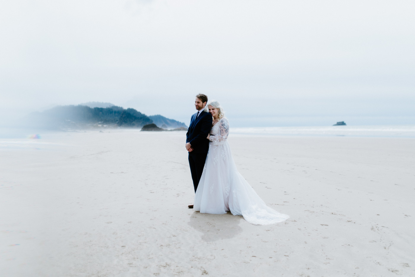 Ben and Katie stand on the sand on the beach along the Oregon coast.