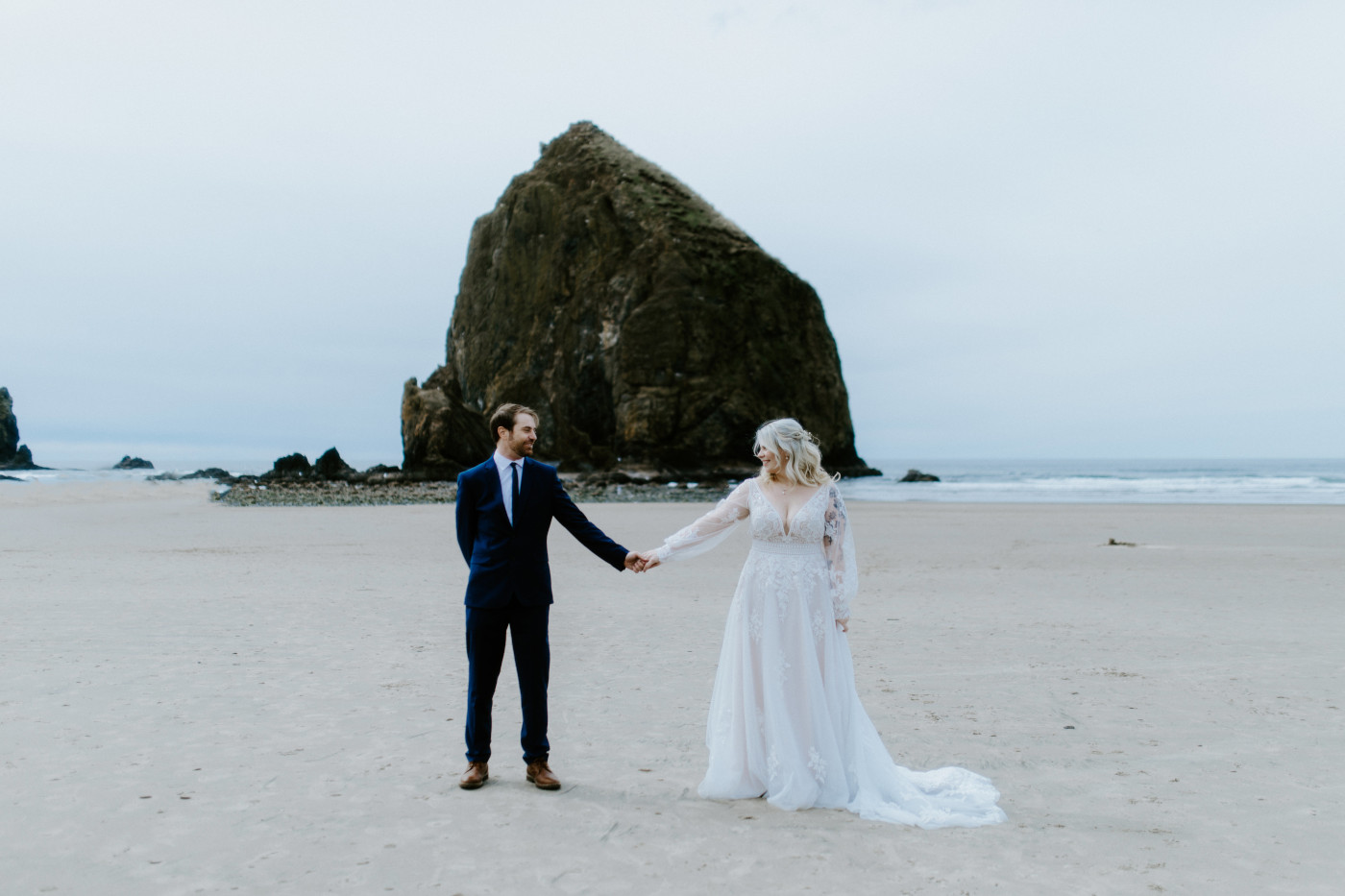 Katie and Ben hold hands on the sand on the beach along the Oregon coast.