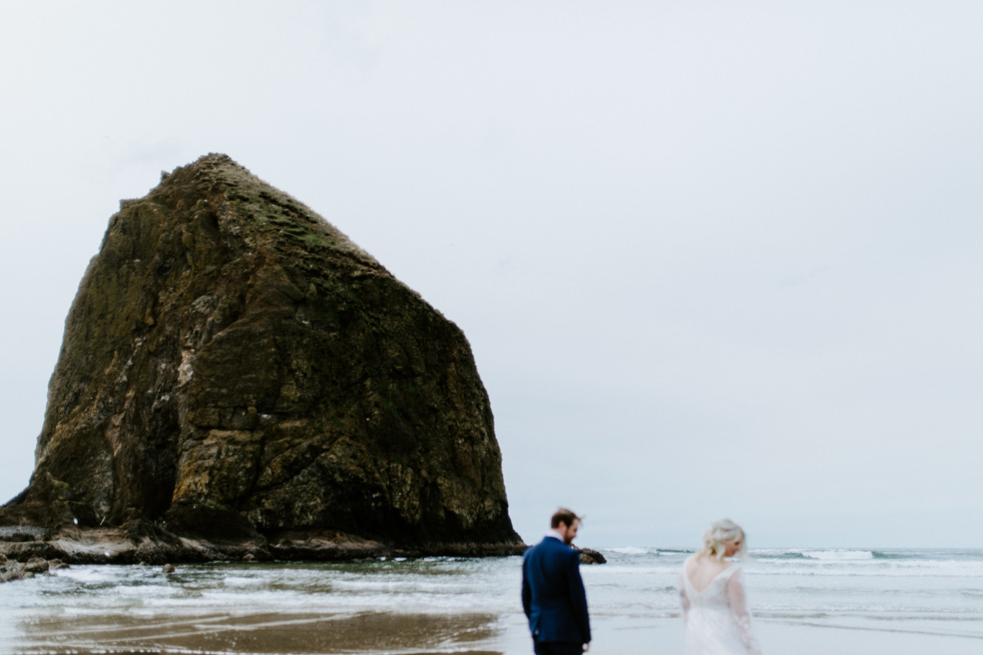 Katie and Ben stand on the sand with a big rock on the Oregon coast.