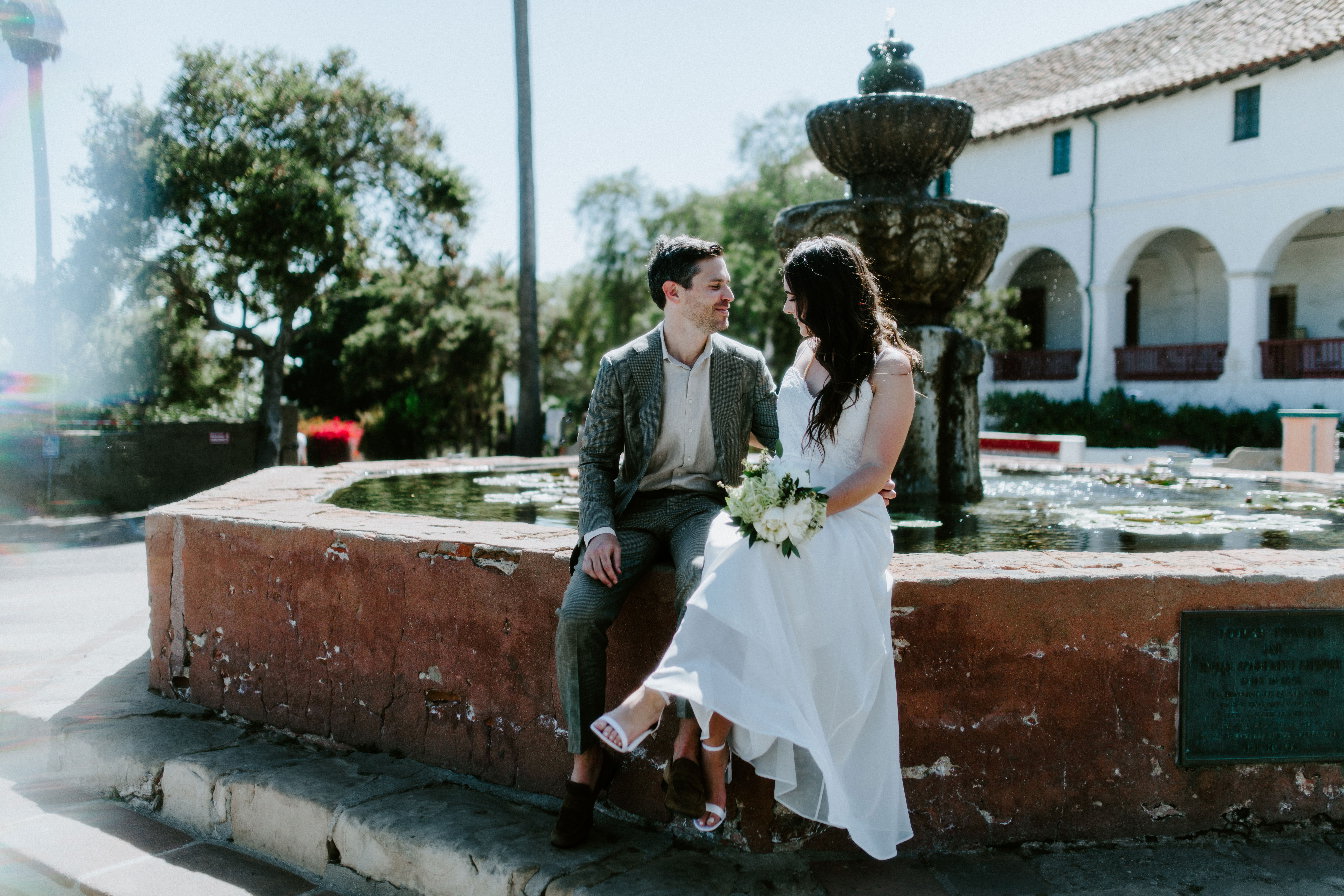 Newlywed elopement couple sits on a historic fountain outside of the Santa Barbara Mission in Santa Barbara, California.